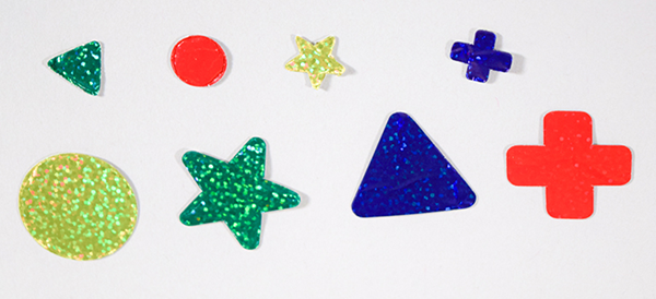 Photograph of a sampling of red, blue, green, and yellow precut holographic adhesive paper stickers showing large and small circles, stars, plus signs, and triangles.