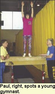A girl stands on a pommel horse. Paul and an assistant stand on either side.