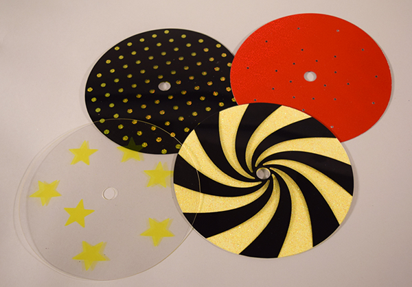 Photograph of four overlays with mid-level complexity: a red holographic overlay with perforations, black and clear dot overlay, clear overlay with yellow stars, and black pinwheel overlay on top of a yellow sparkle overlay.