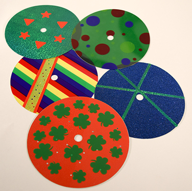 Photograph of five overlays with high complexity patterns: multi-color stripe overlay modified with yellow and red sticker strips; blue holographic overlay modified with green sticker strips; multicolor dot overlay on top of green holographic overlay; green holographic overlay modified with red heart and star stickers; and clear overlays with green shamrock patterns on top of red holographic overlay.