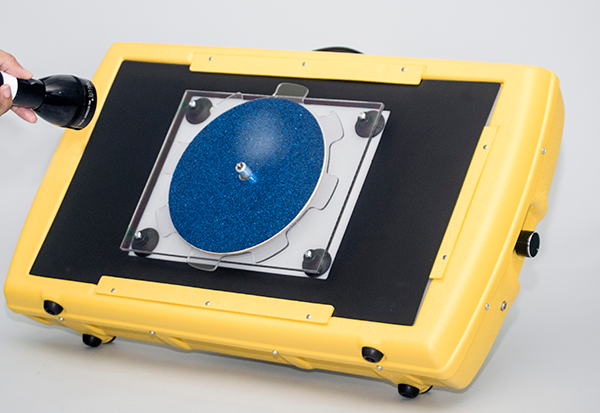 Photograph of the APH Light Box set up for use with the rectangular blackout sheet from the Level 1 Light Box Materials, the APH Plexiglas® Spinner, and a blue sparkle overlay. A flashlight is shown shining light on and reflecting off the overlay.