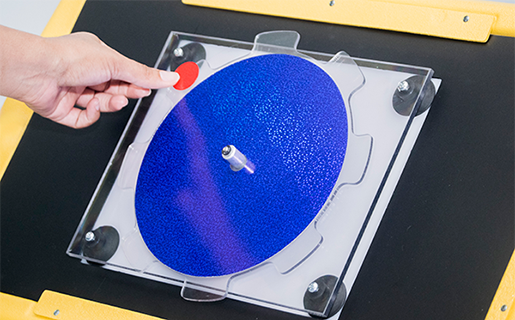 Photograph of the APH Light Box set up for use with the rectangular blackout sheet from the Level 1 Light Box Materials, the APH Plexiglas® Spinner, and a blue sparkle overlay. As a visual location cue, the edge of the spinner is highlighted by a large red sticker placed at eleven o’clock.