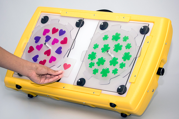 Photograph of the APH Light Box and two APH Plexiglas® Spinners each with different patterned overlays (hearts and shamrocks). The photograph demonstrates the use of a Supplemental card with a heart which matches the hearts on one of the overlays.