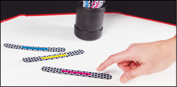 A black plastic cylindrical cup in front of a black felt tri-fold board holds several Phase II Advanced MATCH STICKS. Three sticks lie on a white surface next to the cup; a hand points to a MATCH STICK with a pink stripe.