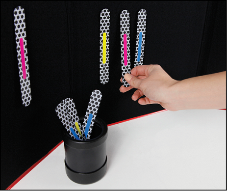 A black felt tri-fold board with four attached Phase II Advanced MATCH STICKS: one pink-striped stick is set apart from a group of yellow-, pink-, and blue-striped sticks. A hand pulls the pink-striped MATCH STICK away from the group. A black plastic cylindrical cup in front of the board holds several more Phase II Advanced MATCH STICKS.