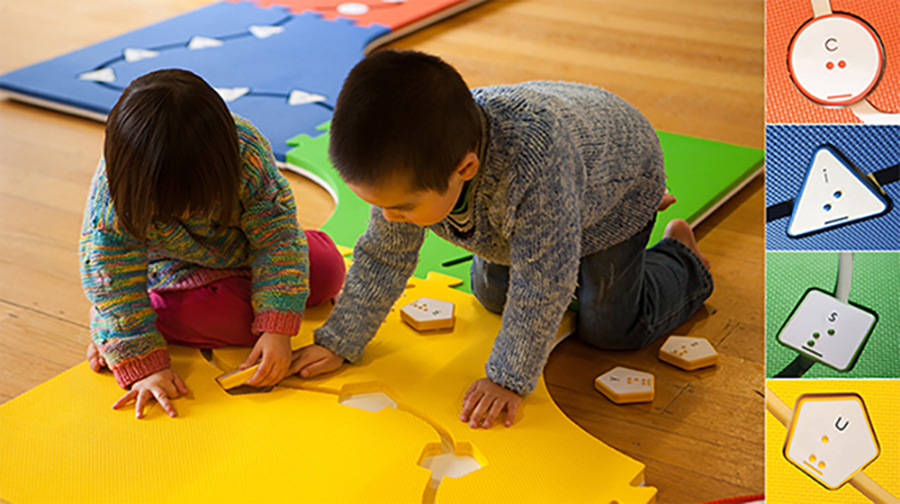 Two students fit Reach & Match sensory tiles into pockets of the Reach & Match sensory play mat. On the right side, there are four pictures of sensory tiles. Print and braille letters are on the tiles as follows: c on a red circle tile, i on a blue triangle tile, s on a green square tile, and u on a yellow pentagon tile.