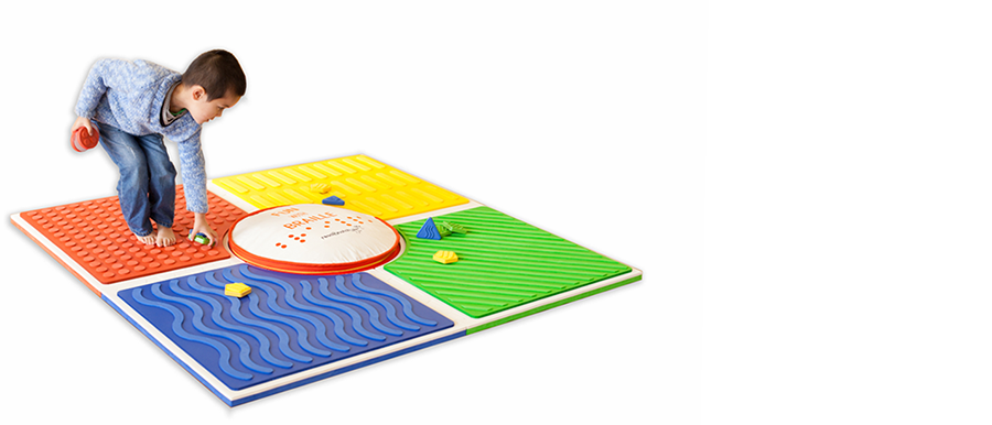 A boy stands on the four connected Reach & Match sensory play mats; he picks up sensory tiles that are scattered across the mats. The Big Round Cushion is in the center of the play mats.
