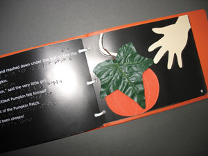 A tactile illustration is shown that combines a large foam paper cutout of a pumpkin, a raised line drawing of a child's hand, and a large silk leaf.