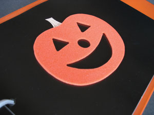 A jack-o-lantern shape cut from thick foam paper and glued to a highly contrasting, black paper page.