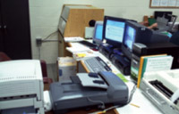 Desk littered with three flickering computer monitors, a copier, scanner, and paperwork.