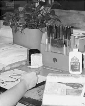 A tactile graphic is being created. Next to the graphic and the artist's hand are the tools and supplies needed, and the book with the original illustration.