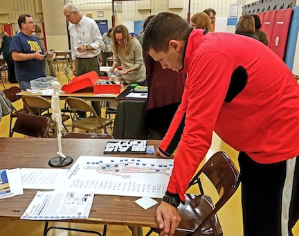 A man in a red jacket examines the APH Touch, Label, and Learn Human Skeleton. In the background, other APH products are displayed