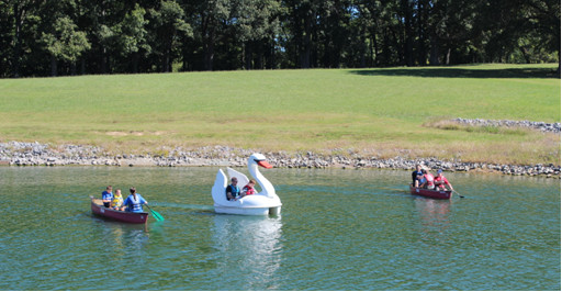 Students and adults paddle two canoes and a swan boat on a lake.
