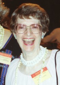 Verna Hart, Color photograph of woman, Verna Hart, with short brown hair and a big smile on her face. She wears large eyeglasses, a white dress, a bead necklace, and a name badge.