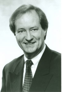 Black-and-white, head-and-shoulders photo of Tuck Tinsley. He is looking at the camera and smiling; dark suit jacket, light-color dress shirt and patterned tie; short hair.